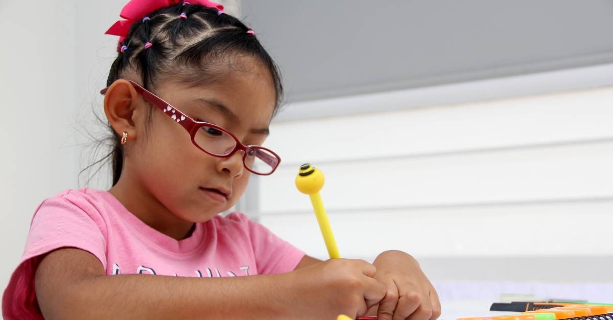 A little girl wearing pink glasses sits at a table and writes with a yellow pen.