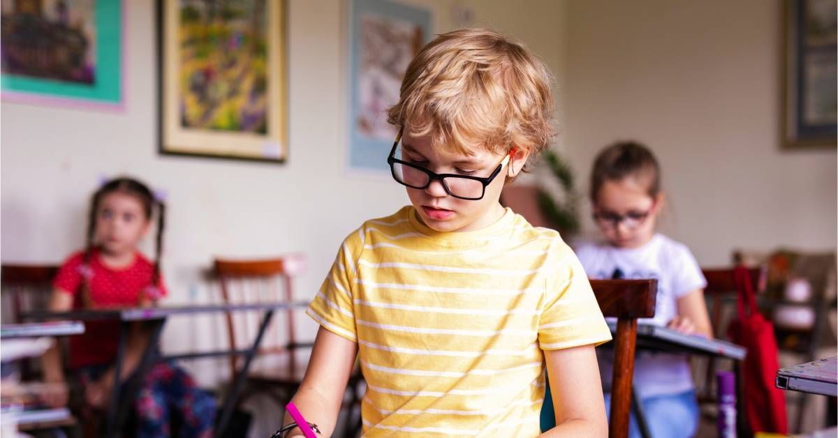 A little boy wearing glasses uses a pencil to draw on a piece of paper in class.