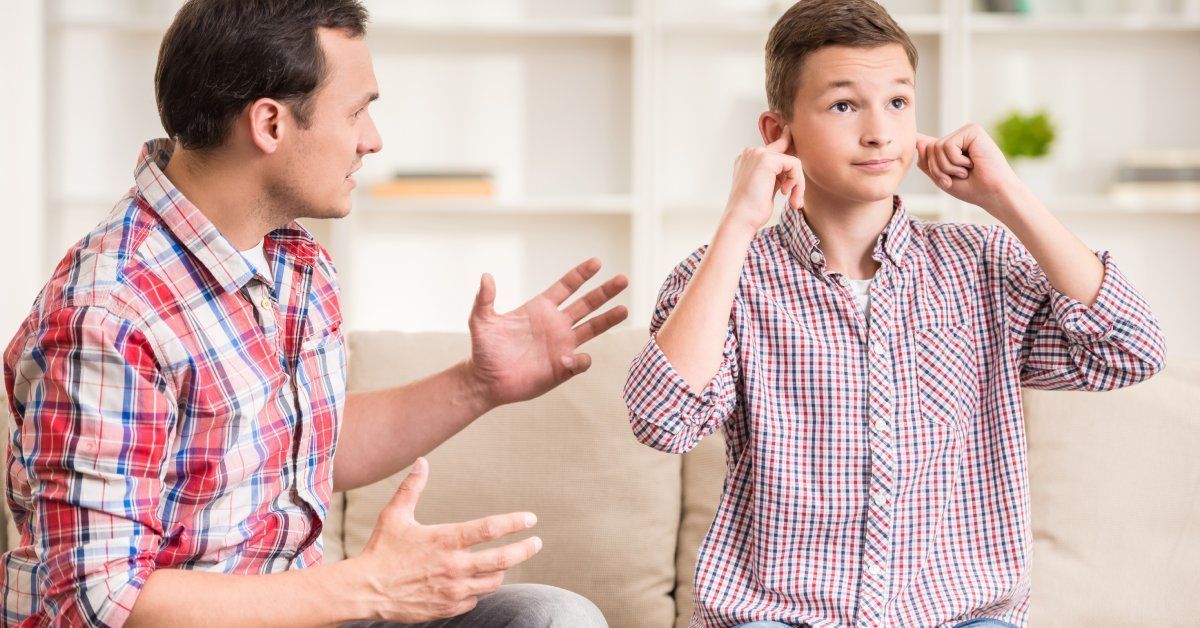 A young boy sitting on a couch while plugging both ears with his fingers as he sits next to an older man.