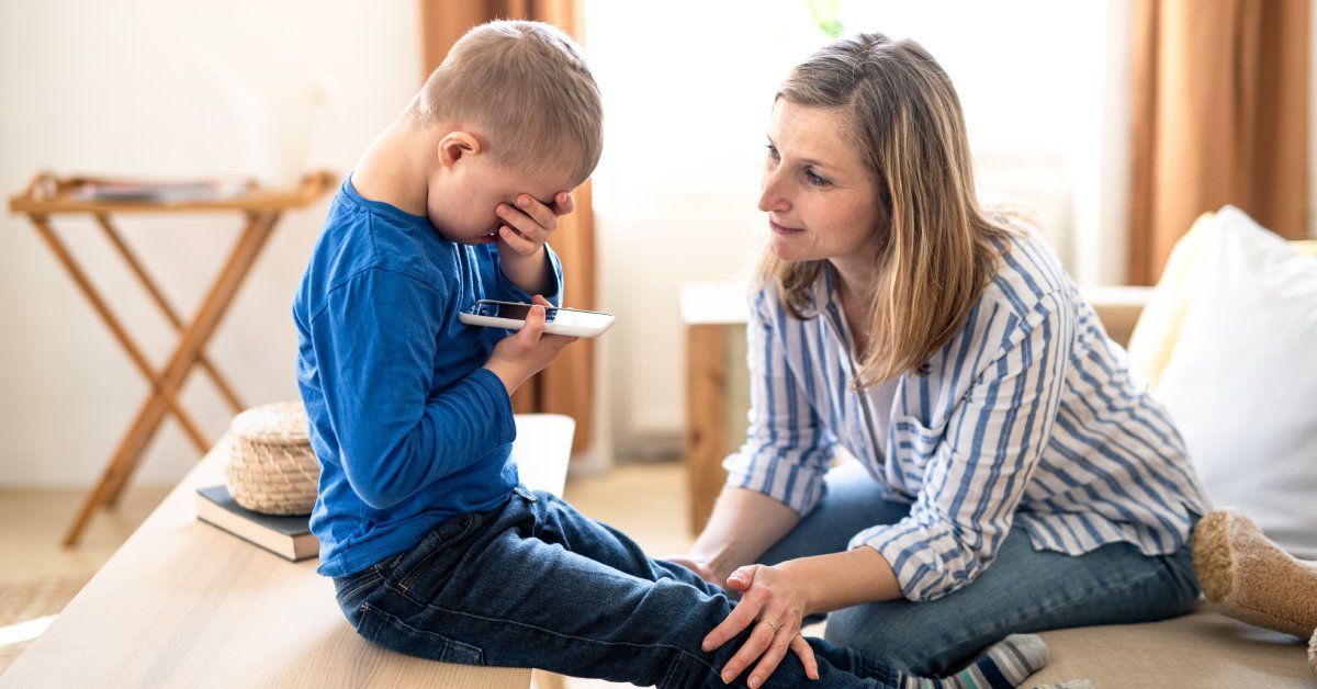 A young boy sitting with his head in his hand while holding a phone. An older woman is kneeling in front of the boy trying to comfort him.