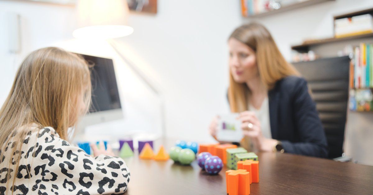 A woman sits at a desk across from a girl. Colorful blocks are on the desk.