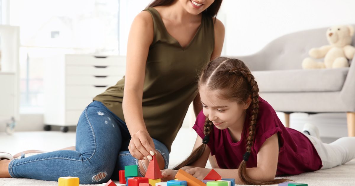 A young woman wearing green. She sits on the ground next to a little girl who plays with a set of multi-colored blocks.