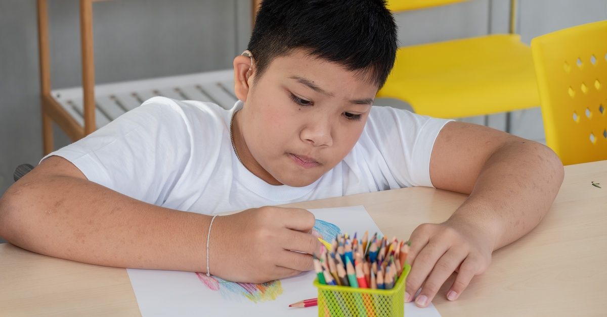 A child sits at a desk while using colored pencils to color on a piece of paper.