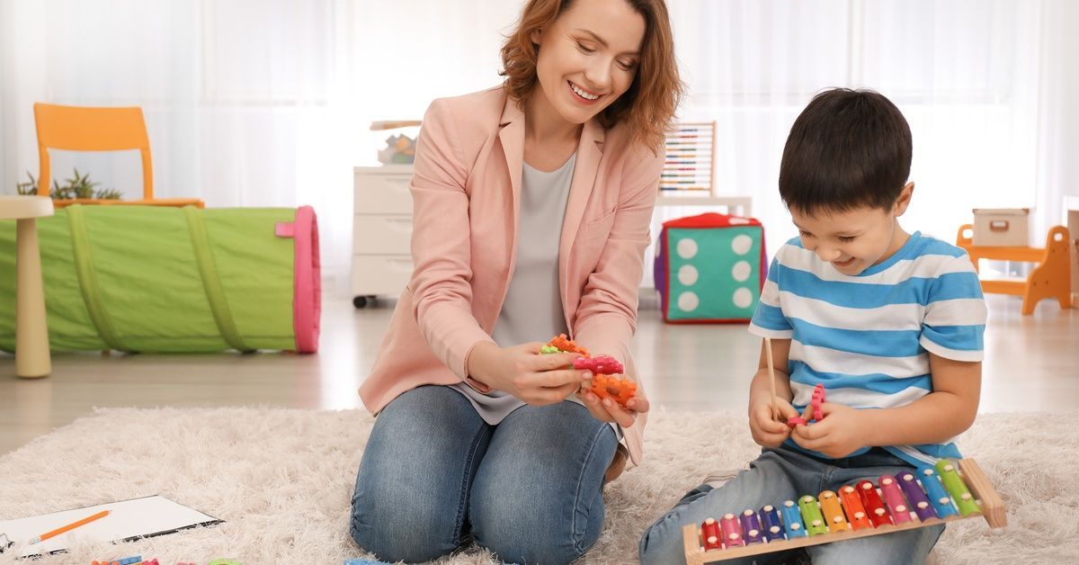 A little boy wearing a striped shirt sits on the ground next to a woman while holding a xylophone in his lap.