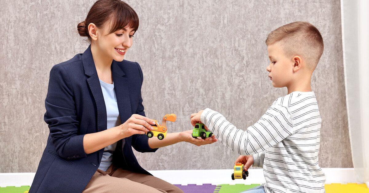 A little boy wearing a striped shirt sits on his knees while handing a toy car to a woman sitting next to him.