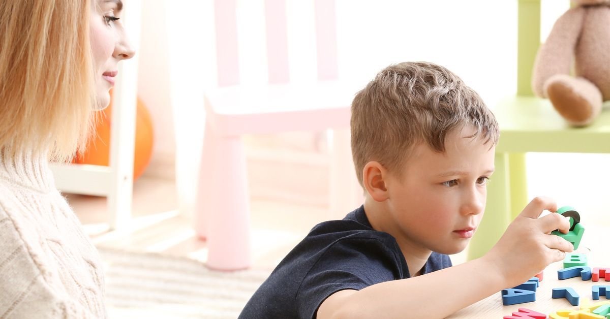 A little boy wearing a blue shirt sits at a table while playing with alphabet tiles. A woman sits next to him while writing.