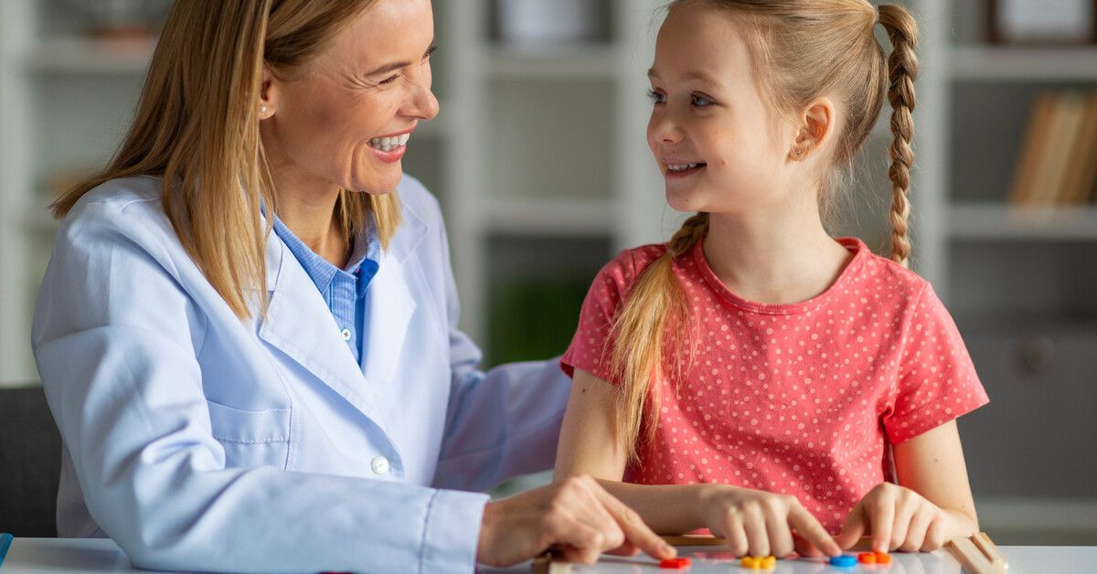 A woman wearing a white coat sits at a table next to a smiling little girl who is playing with alpha