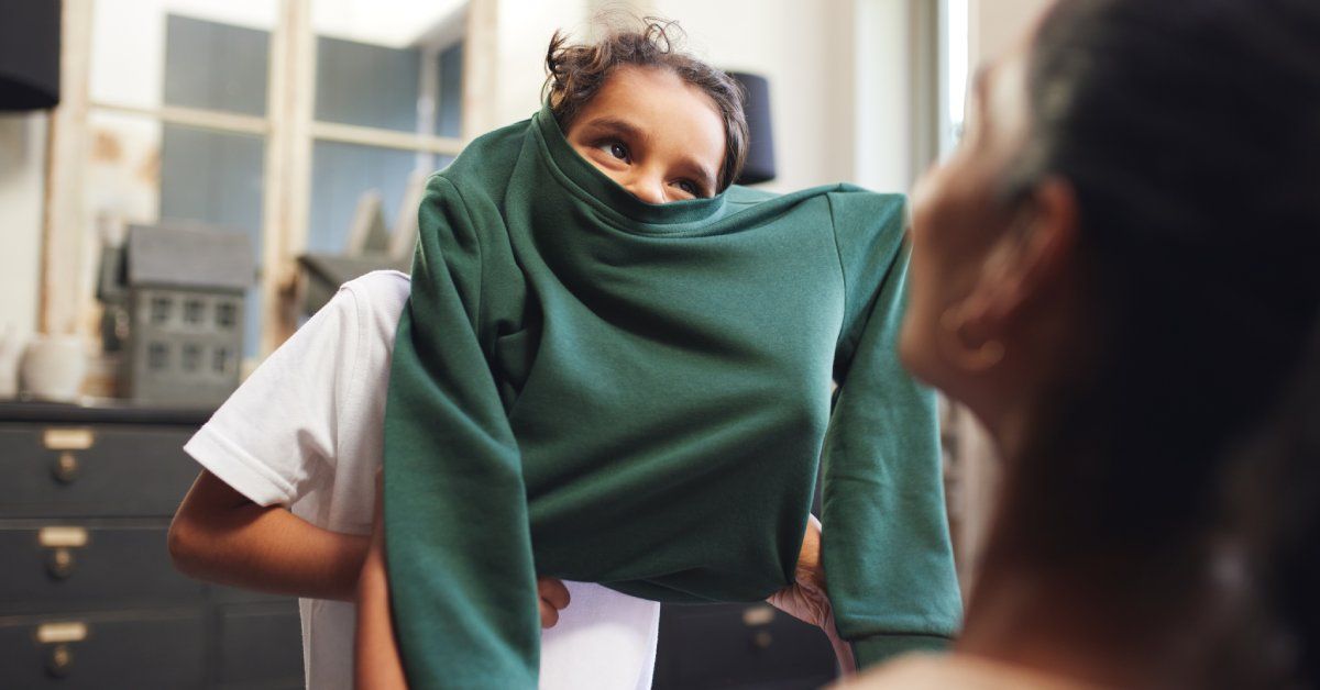 A little boy stands in front of a woman, who helps him to pull a green sweatshirt over his head. They stand in a bathroom.