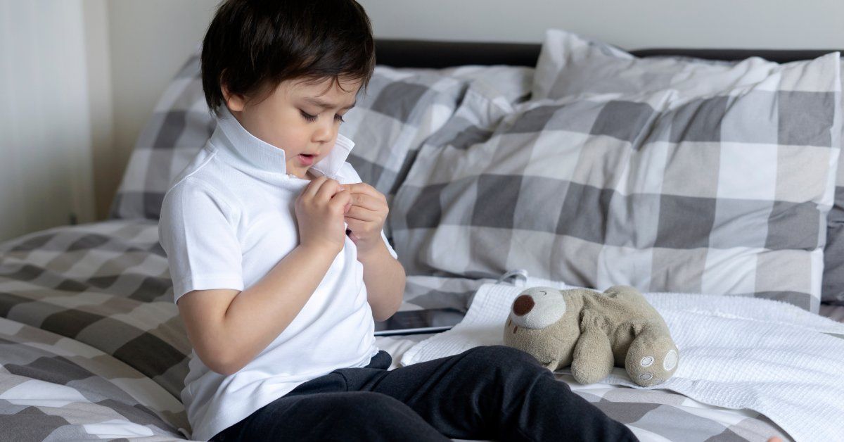 A little boy sets on the edge of a bed while trying to button his white shirt.