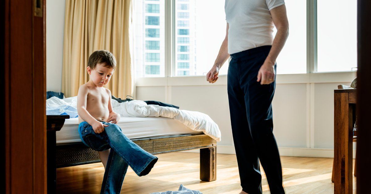 A man wearing a white shirt stands near a young boy who sits on the edge of a bed while putting on pants.
