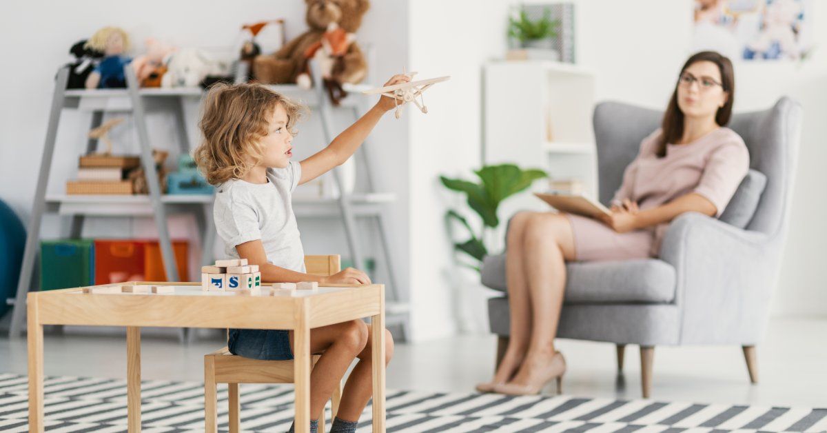 A boy sits at a desk while playing with a toy airplane while a woman watches.