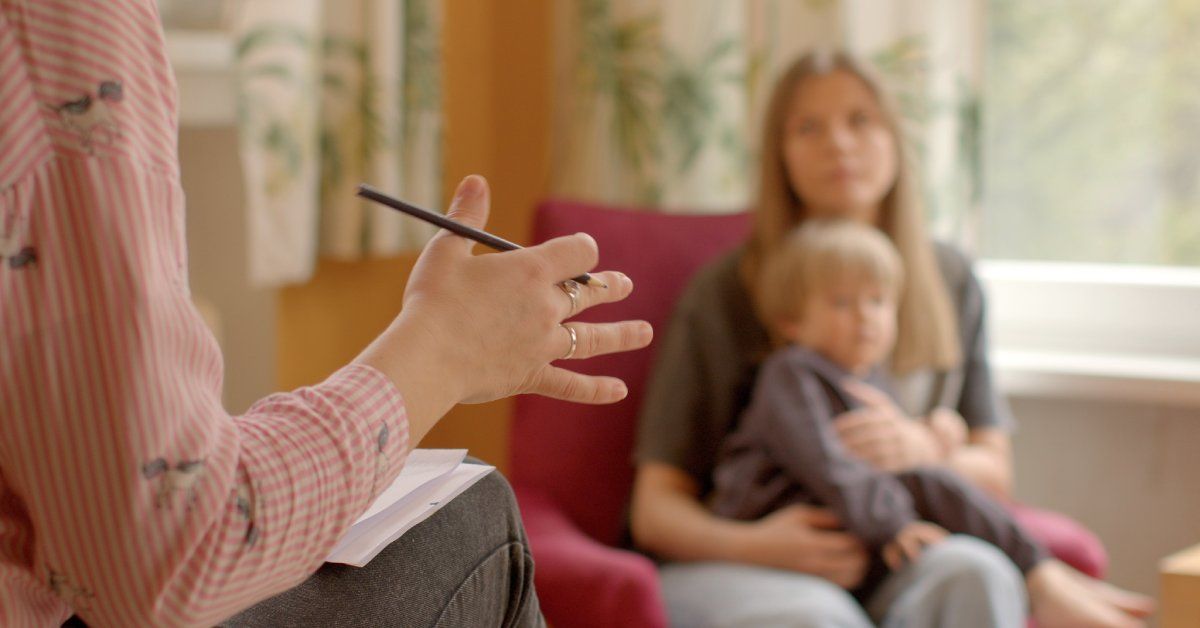 A woman gestures with her hands while holding a pen and sitting across from a woman with a child in her lap.