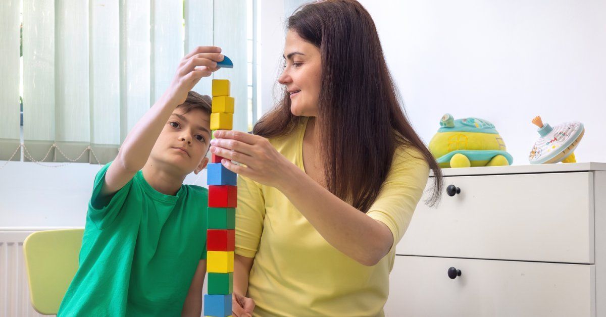 A little boy sits at a table next to a woman while stacking multicolored building blocks on top of one another.