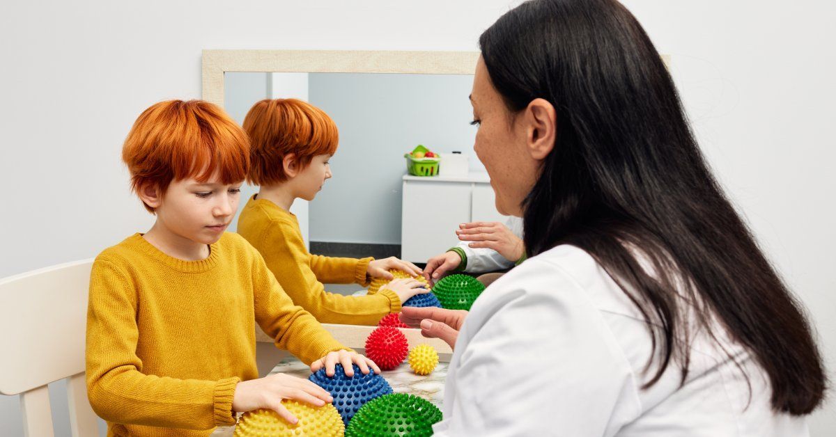 A little boy wearing a yellow shirt sits at a desk across from a woman while touching sensory objects in several colors.