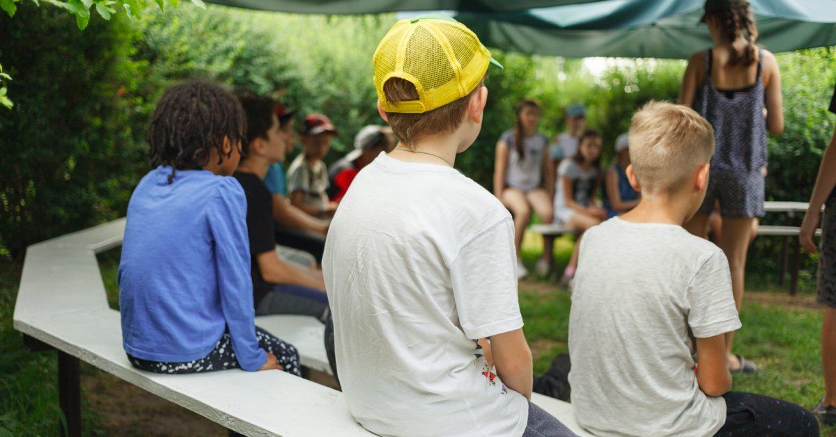 A group of young children sitting on rows of benches under a blue-colored canopy while a young woman stands in the middle.