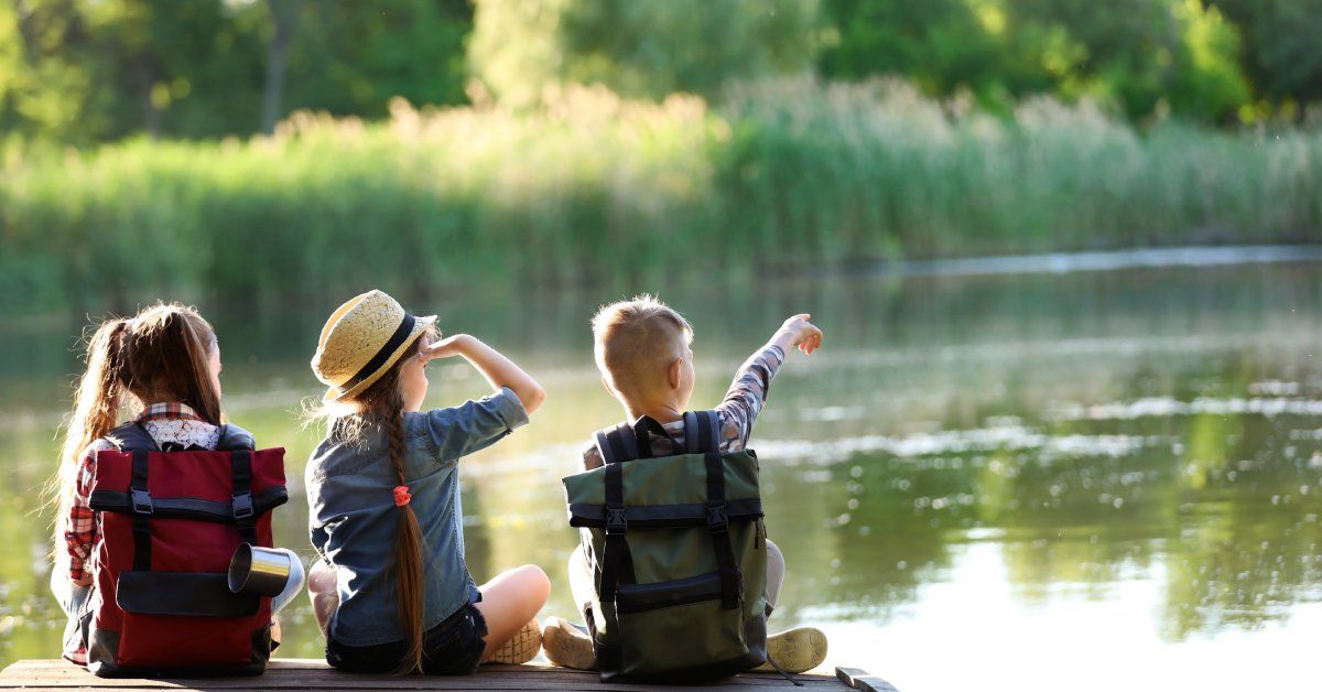 Three young children sit on a dock overlooking a lake surrounded by trees.