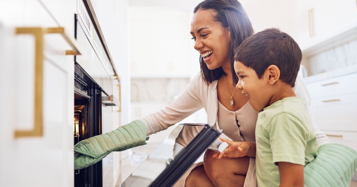 A young woman kneels down to open an oven next to a little boy wearing a green shirt. Both smile while looking at the oven.