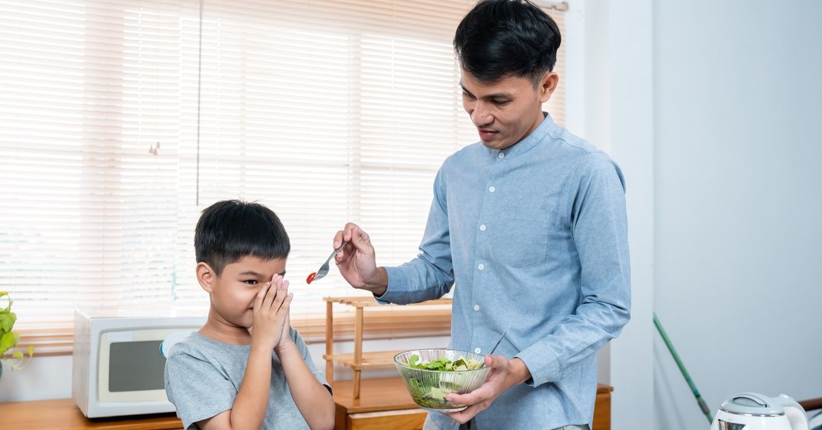 A man stands next to a young boy at a table while trying to feed him. The child holds both hands to his face.
