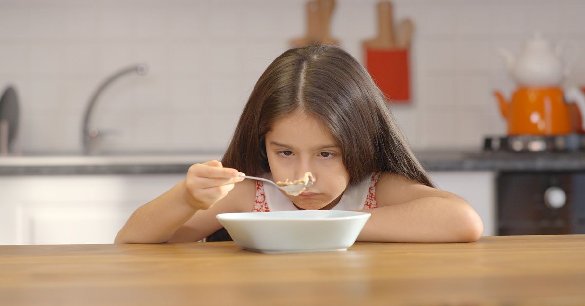 A little girl sits at a kitchen table while staring at the food in her spoon.