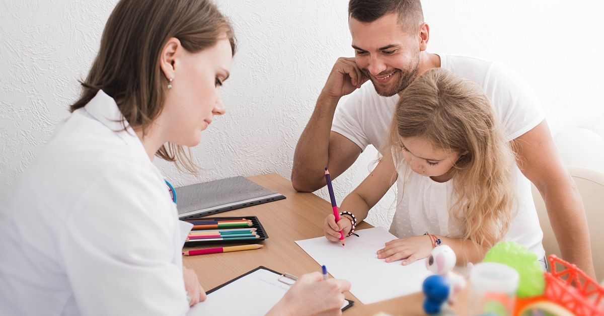 A man sits at a table with a little girl in his lap as she uses a pencil to color a piece of paper. A woman sits across from them.