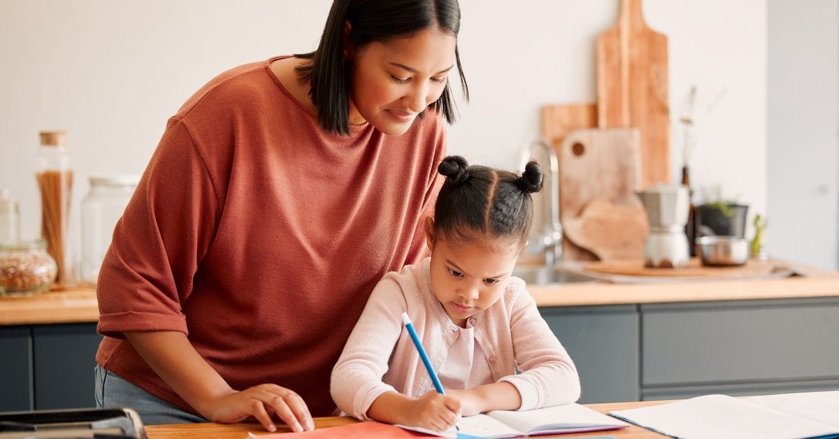 A woman stands over a little girl who's sitting at a table while using a pen to write on a piece of paper.