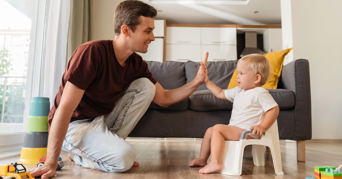 A little boy sits on a small potty on a wood floor. A man smiles at the boy while giving him a high five.
