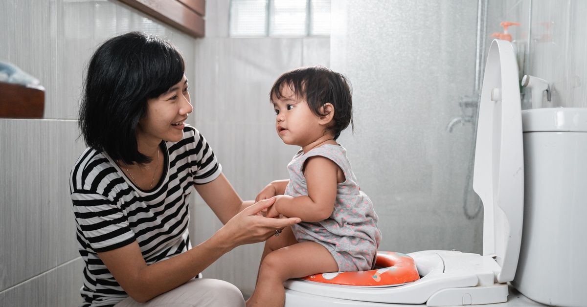 A woman kneels down next to and holds the hands of a small toddler who sits on a potty seat on a toilet.