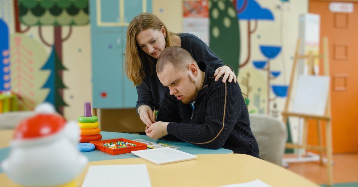 A woman stands over the shoulder of a young man who sits at a table while looking at a box of puzzle pieces.