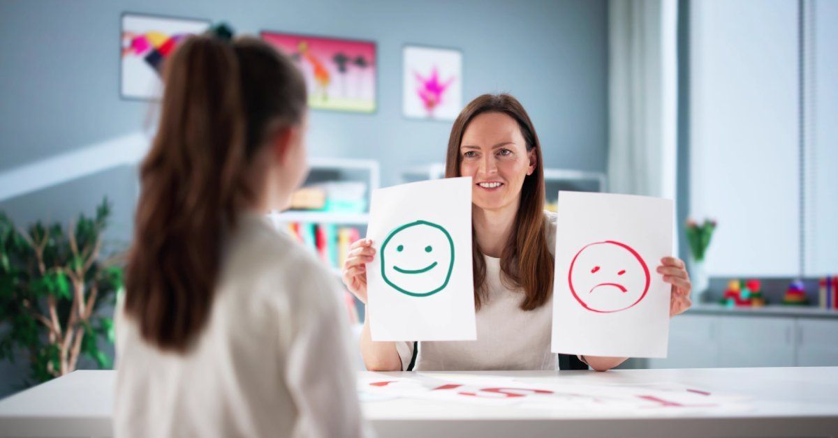 A woman holds a piece of paper with a smiling face, and the other with a frowning face while sitting across from a girl.