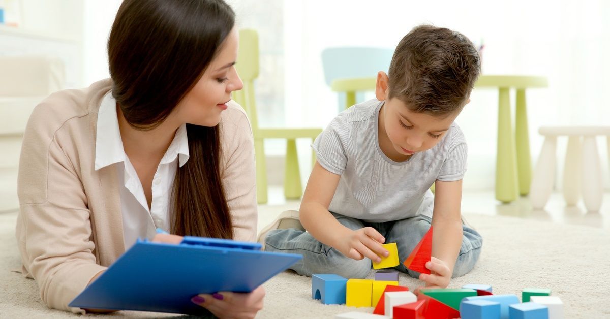 A child plays with blocks on the ground. A woman with a clipboard sits nearby.