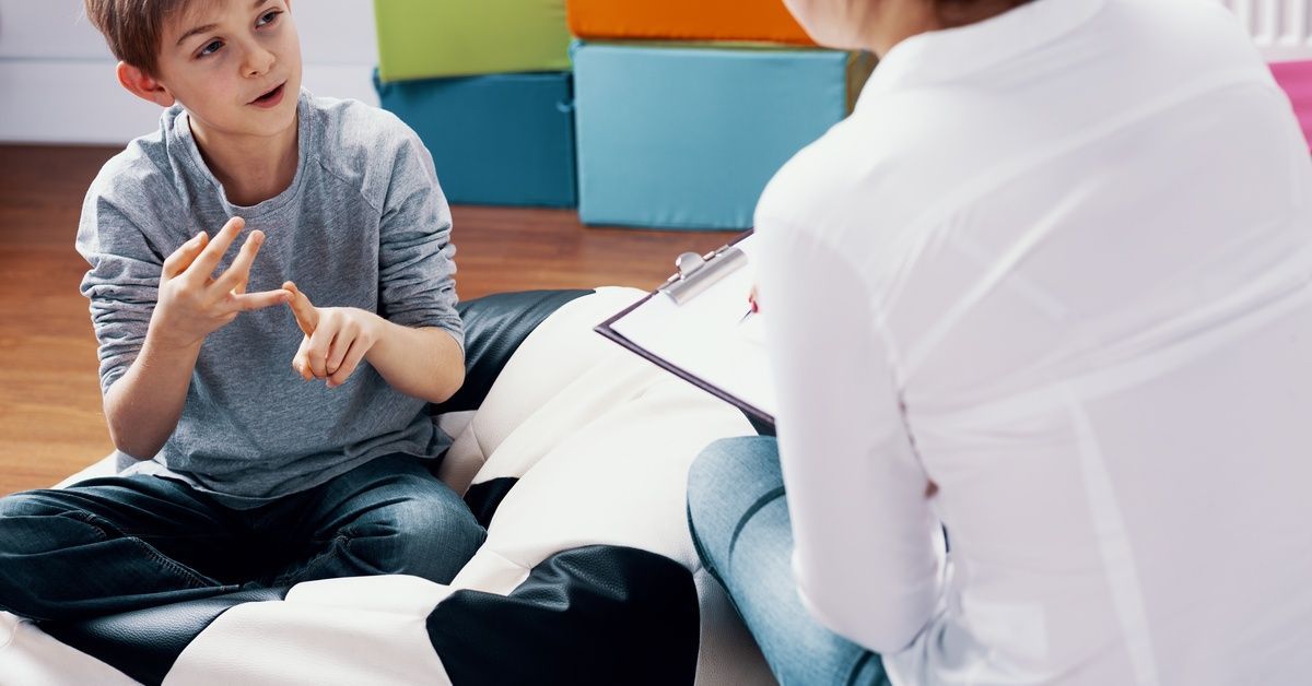 A little boy sits on a soccer ball beanbag while talking to the woman sitting across from him. She holds a clipboard.