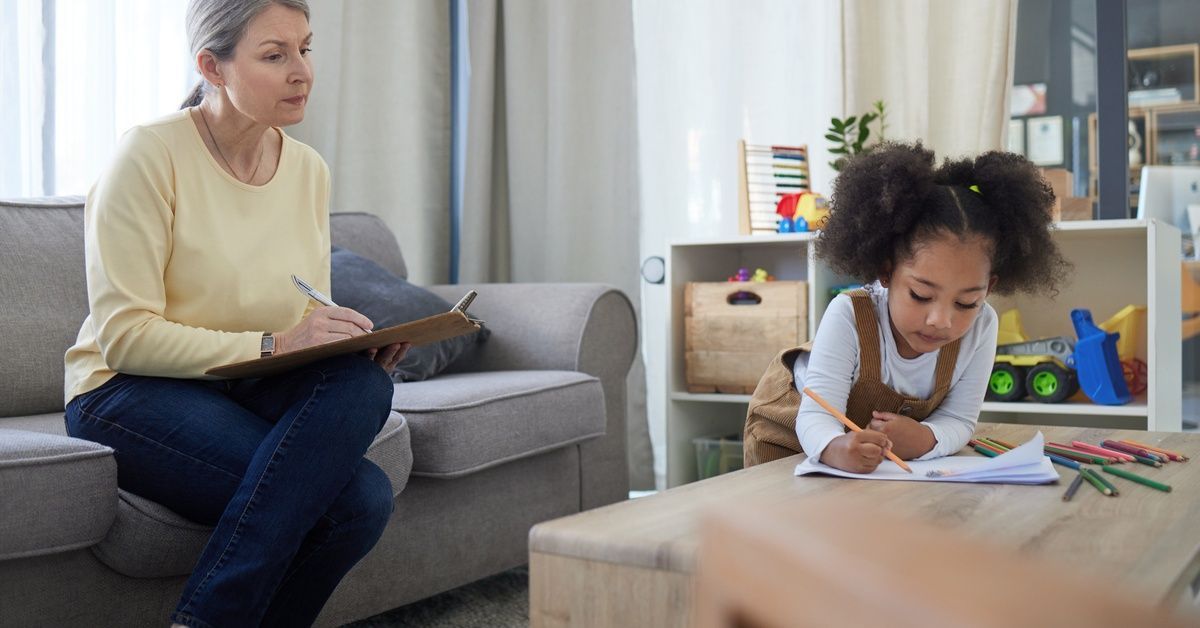 A woman sits on a couch while holding a pen and clipboard. She observes a little girl playing in front of her.