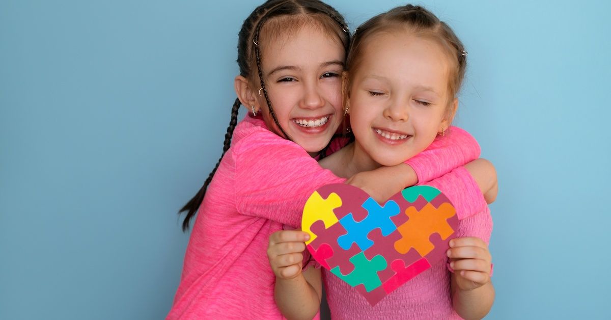 Two girls, both wearing pink, smile and hug while holding up a heart made out of puzzle pieces.