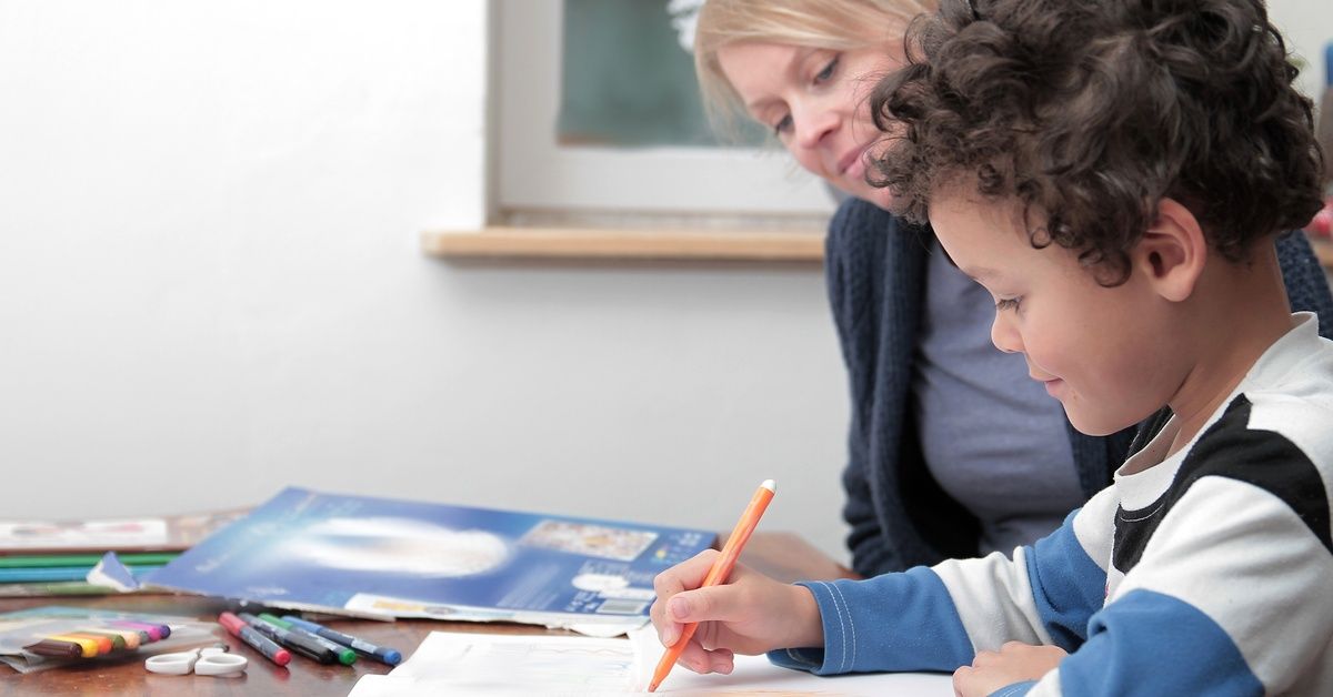 A little boy wearing a striped shirt and sitting at a table while using a marker to color a piece of paper. A woman is sitting next to the boy as she watches him write.