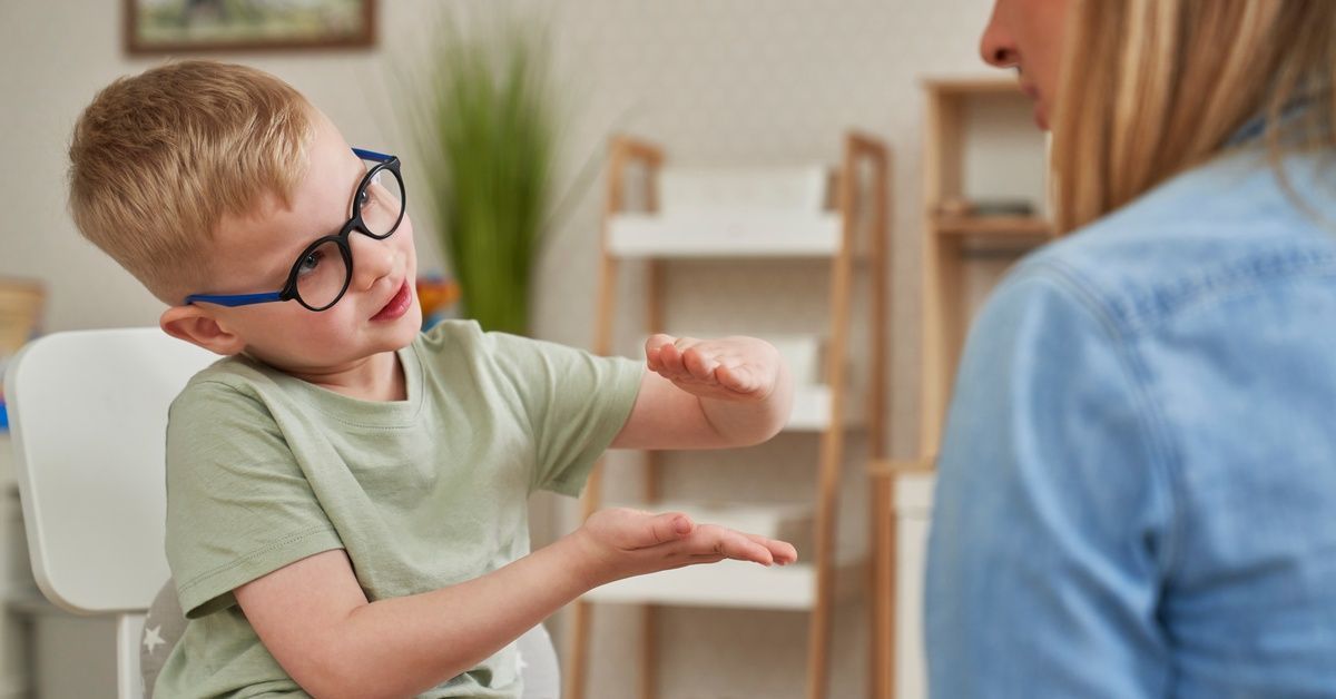A little boy wearing round glasses sits in a chair while gesturing toward the woman sitting across from him.