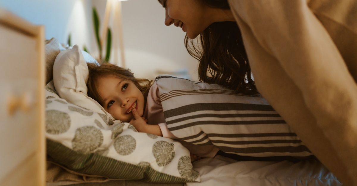 A young woman smiling while leaning toward a little girl, who smiles while laying under the covers in bed.