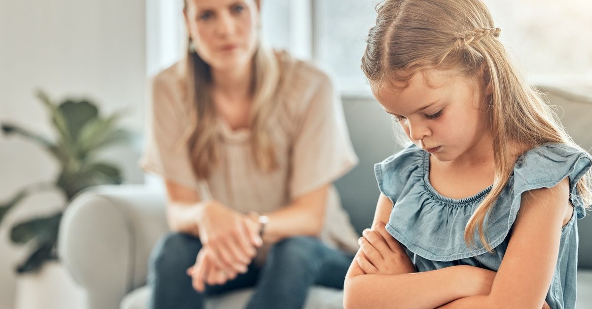 An upset looking little girl sits with both her arms crossed while looking down. A woman sits in the background.