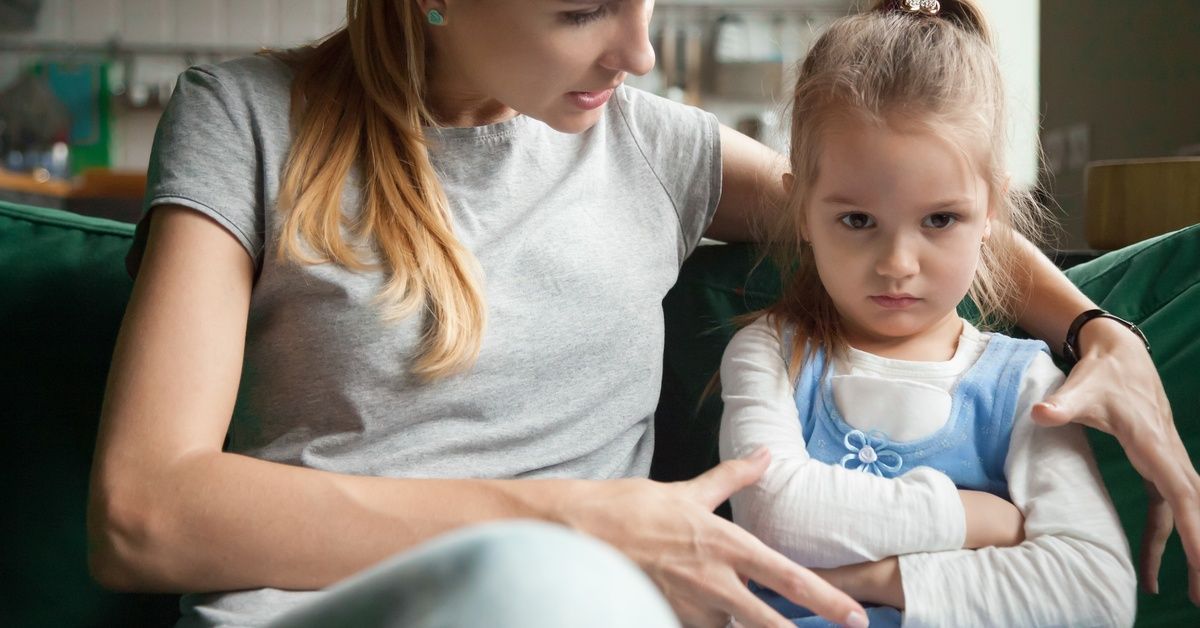 A woman and an upset-looking girl sit together. The little girl has her arms crossed while the woman speaks to her.