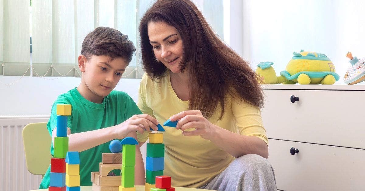 A young boy wearing a green shirt sits at a table while playing with building blocks. A woman sits next to him.