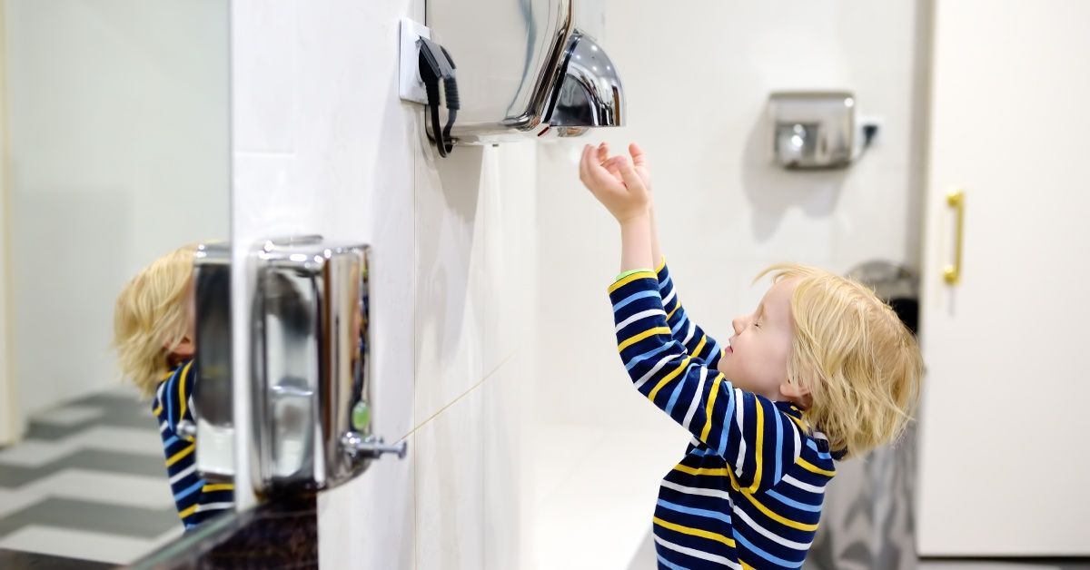 A little boy wearing a striped shirt holding his hands up to a hand air drying machine in a public restroom.