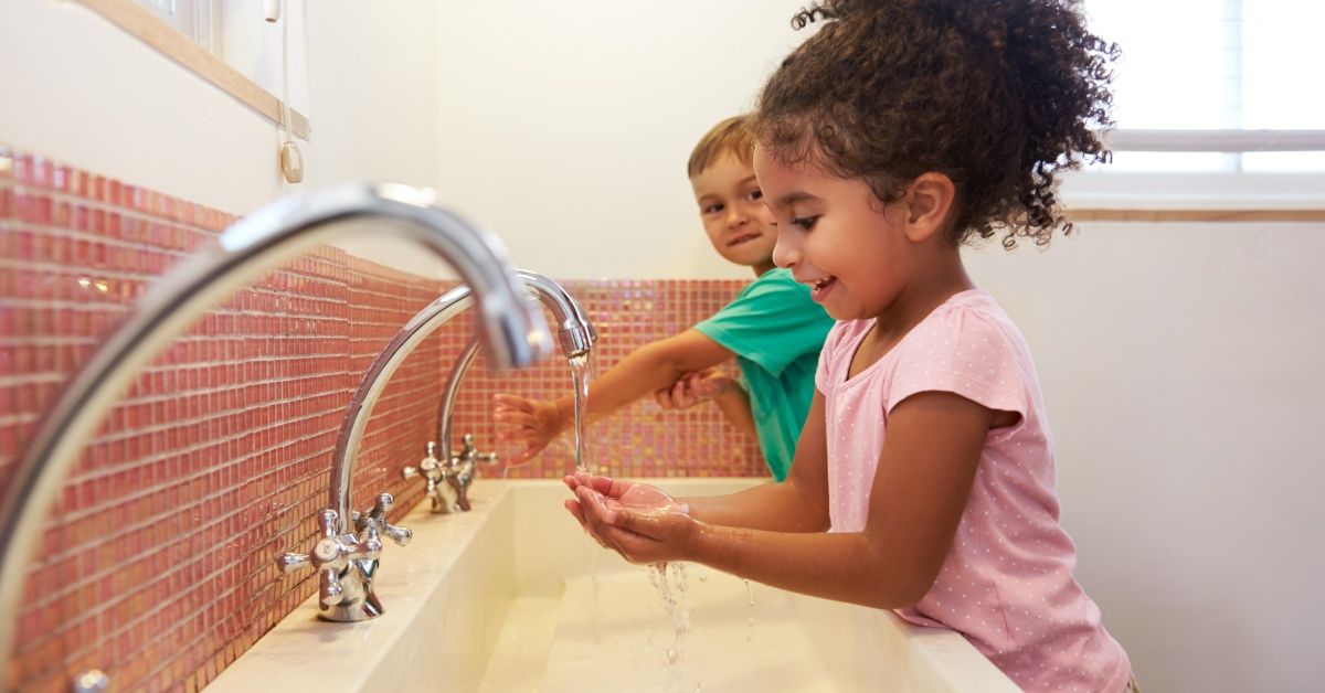A boy washing his hands while looking over at a little girl washing her hands.