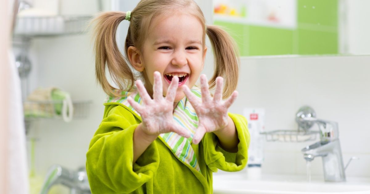 A little girl wearing a bright green-colored robe standing near a sink and smiling while holding up soapy hands.