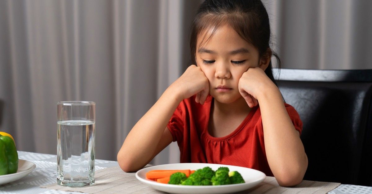 A little girl rests her head on her hands while staring at her plate of veggies.