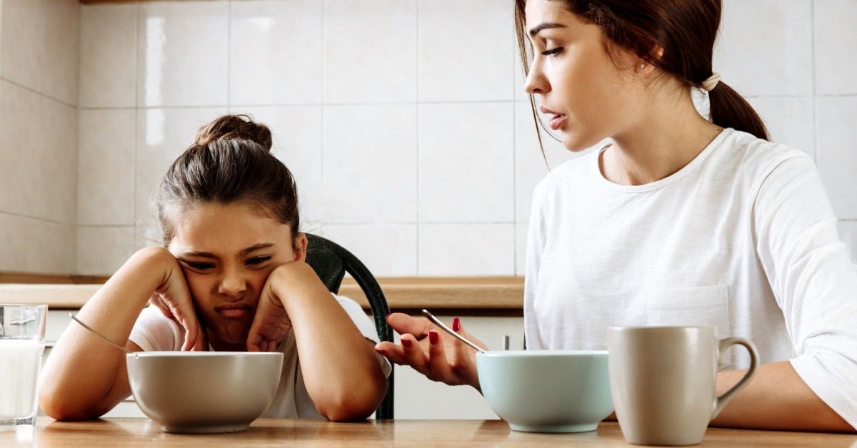 A woman sits next to a young child at a kitchen table. The child frowns at the food while the woman speaks to her.