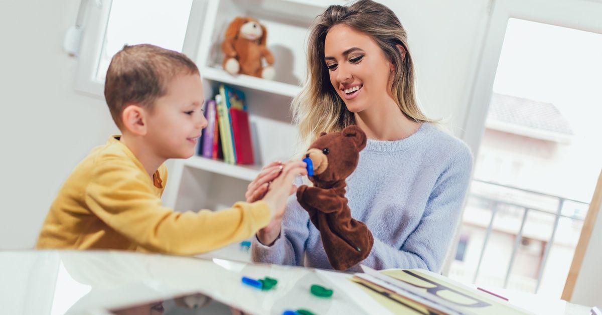 A little boy smiles while sitting next to a woman with a teddy bear sock puppet.