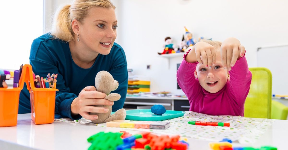 A little girl wearing a purple shirt smiles and holds up her hands while sitting next to a woman at a table.