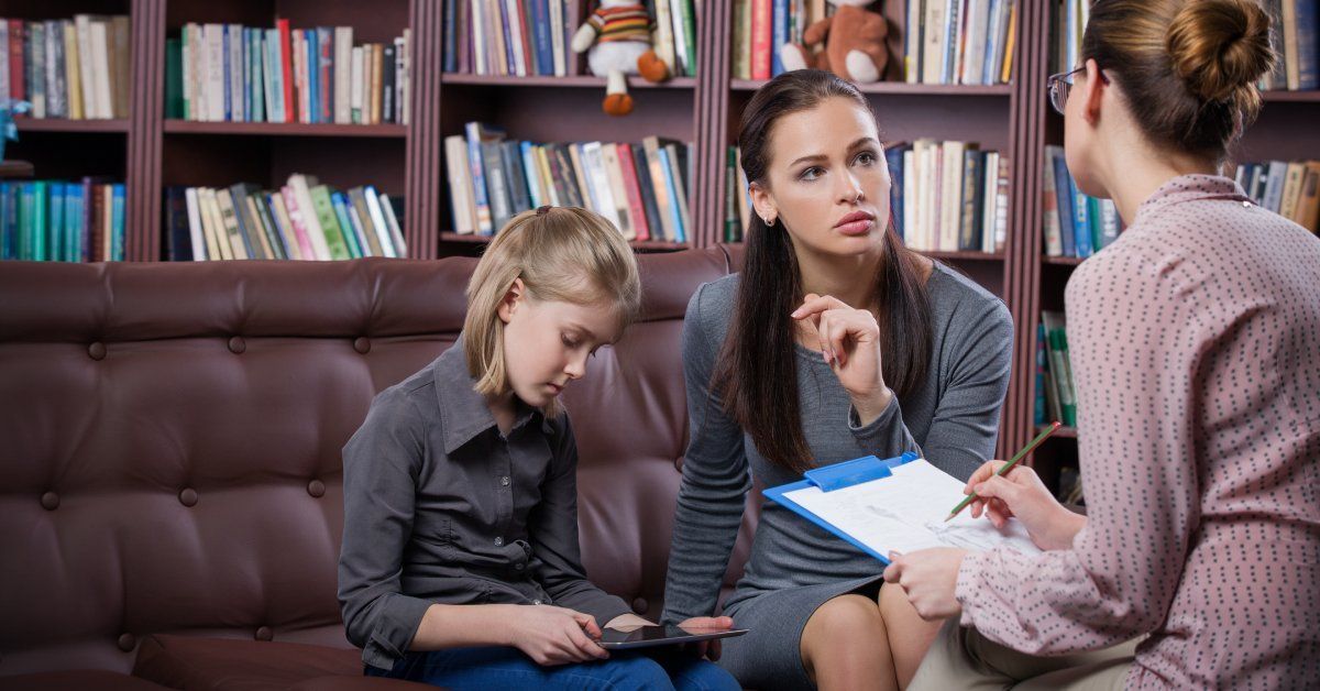 A young woman sitting on a couch next to a little girl while speaking to a woman across from her in a library.