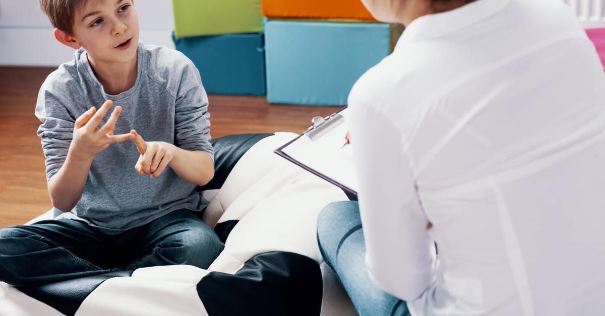 A young boy wearing a gray shirt sits on a beanbag near a woman in a chair, while gesturing with both his hands.