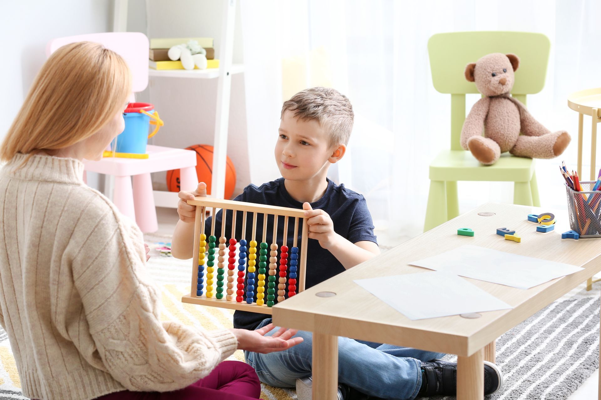 A young boy sitting on the ground next to a table, while holding up a puzzle game toward the woman next to him.