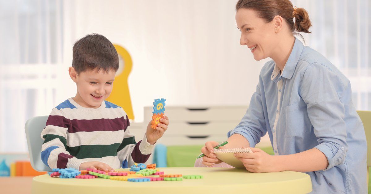 A little boy wearing a striped shirt sitting at a table while smiling and playing with multiple puzzle pieces.